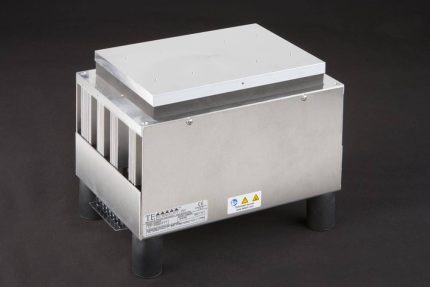 Three-quarter view of large thermoelectric cold plate cooler/heater incorporating six thermoelectric modules. The cooled aluminum plate on the front and waste heat is exhausted to high-density fins on the back. Fins are fan cooled. The fan air enters on the middle back and exhausts on sides. The cooler incorporates legs which lift the fan's air inlet off of a table top, so the cooler can be operated directly on a table. It operates on 24 volts DC and removes 204 watts of heat at a 0°C temperature difference relative to ambient. Internal extender blocks improve the heat pumping capacity at larger temperature differences. Can heat to 100°C without damaging the cooler.